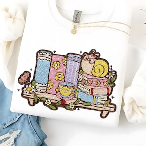 May include: White sweatshirt with an embroidered design featuring a snail wearing glasses, sitting on a stack of books. The design includes a shelf with books, a teacup, flowers, and a butterfly. A gold necklace is visible.
