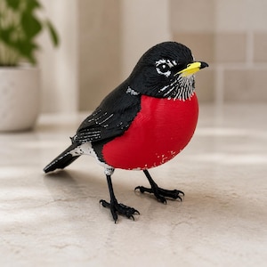 Könnte beinhalten: Eine Vogelfigur mit schwarzem Kopf, Flügeln und Schwanz, einer leuchtend roten Brust sowie weißen Markierungen an Kehle und Bauch. Der Vogel hat einen kleinen gelben Schnabel und schwarze Beine und steht in einer detaillierten Haltung.