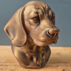 Peut inclure: Buste de chien de couleur bronze avec des détails, notamment des oreilles tombantes et une surface texturée. La sculpture est placée sur un fond neutre, mettant en valeur la tête et le haut de la poitrine du chien.