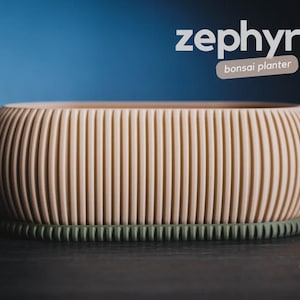 Peut inclure: Un pot à bonsaï beige et nervuré avec une base vert foncé. Le mot "zephyr" est au-dessus, avec "bonsaï planter" en dessous. Le pot est sur une surface sombre avec un fond bleu.