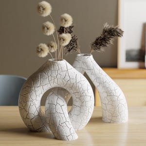 Könnte beinhalten: Zwei weiße Keramikvasen mit Rissstruktur, eine mit Schlaufendesign und die andere mit hoher, geschwungener Form. Beide Vasen enthalten Trockenblumenarrangements. Die Vasen stehen auf einem hellen Holztisch.