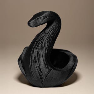 Könnte beinhalten: Eine schwarze, schwanförmige Dekoschale. Die Schale hat eine strukturierte Oberfläche und einen geschwungenen Hals mit einer kleinen Öffnung oben. Der Schwan ist vor einem neutralen Hintergrund platziert, der seine Form und Details hervorhebt.