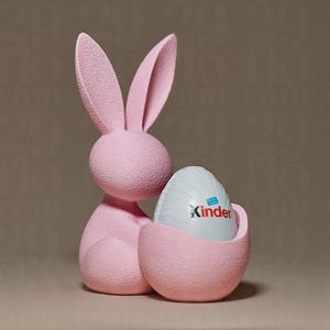 Könnte beinhalten: Eine rosa Hasenfigur, die ein Kinder-Ei hält. Der Hase hat lange Ohren und eine strukturierte Oberfläche. Das Ei ist weiß mit dem Aufdruck "Kinder". Die Figur ist ein dekorativer Gegenstand.