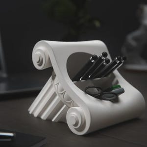 May include: A white, neoclassical-style desk organizer shaped like a stylized column capital. It holds black pens, scissors, and markers. The design features scroll-like volutes and fluted details, suitable for office or home decor.