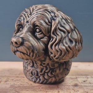 May include: A bronze-colored dog head sculpture with detailed fur texture. The sculpture features a realistic depiction of a dog's face, including eyes, nose, and mouth. The dog's head is mounted on a base, creating a decorative piece.