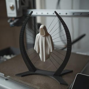 May include: A cream-colored, hooded figure sculpture suspended within a black, arched frame with radial spokes. The sculpture is on a brown surface, with a 3D printer in the background. The artwork has a minimalist, modern aesthetic.