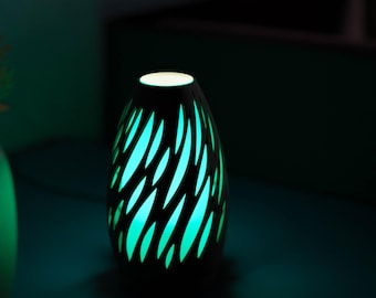 May include: A black, teardrop-shaped essential oil diffuser with a unique cut-out design. The diffuser emits a soft, turquoise glow, creating a relaxing ambiance. The top of the diffuser has a small, white light.