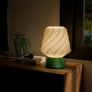 May include: A modern table lamp with a green base and a cream-colored, swirled shade. The lamp sits on a wooden surface, next to a book and two green vases. The lamp's design is contemporary and stylish.