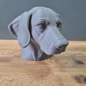 以下が含まれることがあります： 詳細な顔の特徴を持つ、グレーの3Dプリントされた犬の頭の彫刻。犬は長く垂れ下がった耳と目立つ鼻を持っています。彫刻は木製の表面に置かれています。