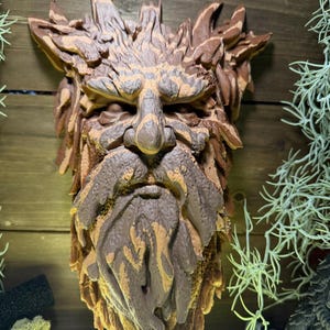 May include: A wall-mounted sculpture of a forest spirit face, crafted with layered brown and tan textures resembling bark and foliage. The figure features a prominent nose, deep-set eyes, and a long, flowing beard made of carved wood-like elements.