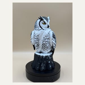 May include: A white and black owl figurine with yellow eyes, perched on a dark tree stump. The owl features detailed feather patterns in black against a white body. The figure is mounted on a circular black rotating display base with visible controls.