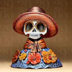 Op de afbeelding: Een Day of the Dead-beeldje met een schedel met geschilderde ogen en een breedgerande hoed. Het figuur draagt een blauwe jurk versierd met oranje en gele bloemen. De hoed en sjaal zijn bruin.
