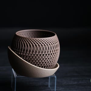 May include: A brown, spherical planter with a woven design, resting on a matching saucer. The planter is made of a dark brown material and sits on a light brown saucer. The design is modern and decorative.
