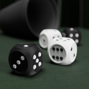 Könnte beinhalten: Ein Set aus drei Würfeln mit abgerundeten Kanten und einer texturierten, geschichteten Oberfläche. Ein Würfel ist schwarz mit weißen Punkten, während zwei Würfel weiß mit schwarzen Punkten sind. Ein schwarzer zylindrischer Becher liegt hinter den Würfeln.