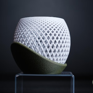 Peut inclure: Objet décoratif sphérique blanc avec un motif en treillis de diamants, reposant sur une base incurvée vert foncé. L'objet est présenté sur un support rectangulaire transparent sur fond sombre. Design moderne et géométrique.