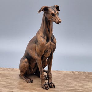 Puede incluir: Una escultura de color bronce de un galgo italiano sentado. La figura presenta una complexión esbelta, orejas puntiagudas y un acabado metálico liso con sombreado oscuro en las hendiduras para resaltar la anatomía muscular del canino.