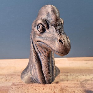 Pode incluir: Um busto de dinossauro na cor bronze com um pescoço longo e uma cabeça detalhada com olhos e narinas proeminentes. A escultura tem uma superfície texturizada com destaques escuros que enfatizam as dobras da pele e os traços faciais.
