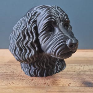 May include: A dark gray dog head sculpture with detailed fur texture. The dog's head is angled slightly to the right, with a focused gaze. The sculpture is on a wooden surface.