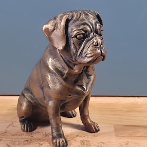 Könnte beinhalten: Eine bronzefarbene Mops-Figur in sitzender Position. Die Skulptur zeichnet sich durch detaillierte Gesichtsfalten, Schlappohren und eine glatte metallische Oberfläche aus. Der Hund ist mit einem konzentrierten Ausdruck dargestellt.