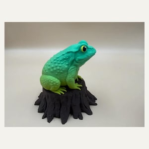 May include: A bright green toad figurine with a textured, bumpy back and large yellow eyes with black pupils. The toad is perched atop a dark brown, cylindrical tree stump base that features vertical ridges and a rough, bark-like surface texture.
