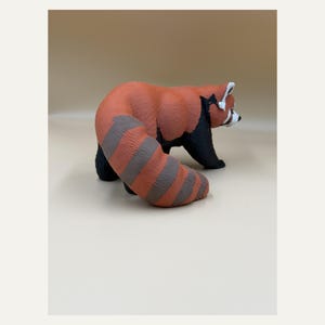 Op de afbeelding: Een rode panda-figuur met een levendige roodoranje vacht, zwarte poten en een zwarte buik. Het dier staat in een staande positie en heeft een lange, dikke, pluizige staart die is voorzien van afwisselende roodoranje en donkerbruine ringen.