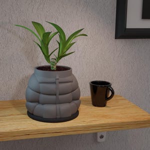 Pode incluir: Um vaso cinza em forma de casaco acolchoado com uma planta verde dentro. Uma pequena caneca preta está em uma prateleira de madeira ao lado do vaso. A prateleira é montada em uma parede texturizada.