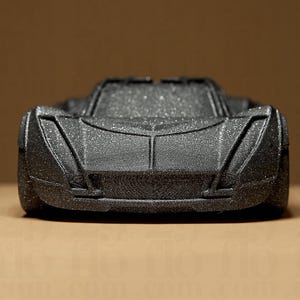 May include: A black toy car with a textured surface and a glittery finish. The car has a low profile, a prominent front grille, and angled headlights. The vehicle is set against a neutral background.