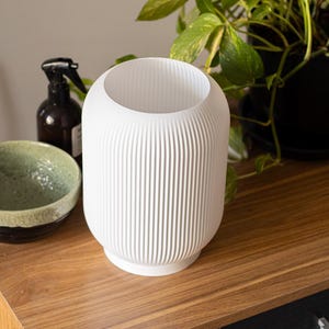 May include: A white, ribbed ceramic vase with a rounded shape. The vase has a wide opening and a textured, vertical stripe design. The vase is sitting on a wooden surface, with a green bowl and a dark brown bottle in the background.
