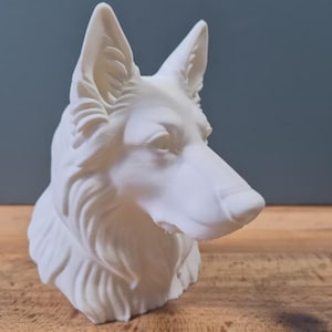 Könnte beinhalten: Eine weiße Skulptur eines Hundekopfes, die detaillierte Merkmale wie Ohren, Schnauze und Fell aufweist. Die Skulptur ist vor einem neutralen Hintergrund platziert, der die Form und Textur des Kunstwerks hervorhebt. Der Hundekopf ist im Profil positioniert.