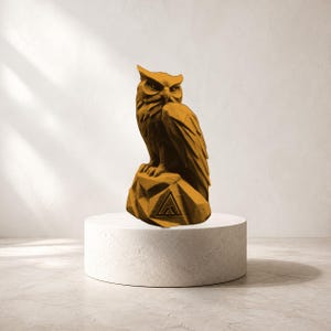 May include: A mustard yellow owl figurine with a geometric, faceted texture perched on a rock base. The base features a triangular emblem with a stylized mountain peak icon engraved on the front, showcasing sharp, angular lines throughout the design.