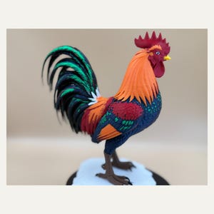 Könnte beinhalten: Eine dreidimensionale Hahn-Figur mit einer lebendigen Farbpalette aus Orange, Rot, Dunkelblau und Grün. Die Skulptur zeigt einen detaillierten roten Kamm, einen gelben Schnabel und lange, fließende Schwanzfedern in Schwarz- und Grüntönen.