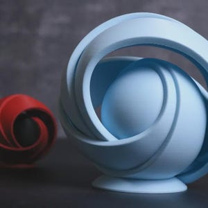 May include: Two abstract sculptures. One is a light blue sphere with a swirling design, and the other is a smaller red sphere with a similar design. Both are on a dark surface.