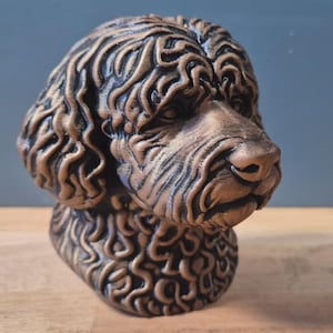 May include: A bronze-coloured dog bust with detailed fur texture. The sculpture features a realistic depiction of a dog's head and shoulders, with a focus on the facial features and curly fur. The bust is displayed on a wooden surface.