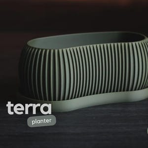 Könnte beinhalten: Ein salbeigrüner Pflanztopf mit geripptem Design, der auf einer passenden Untertasse steht. Das Wort "terra" wird in Weiß angezeigt, darunter "planter". Der Pflanztopf ist ein dekorativer Gegenstand.