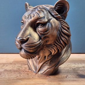Könnte beinhalten: Eine bronzefarbene Tigerkopf-Skulptur mit detaillierten Gesichtszügen, einschließlich texturierter Fellmuster, spitzer Ohren und eines fokussierten Blicks. Die Büste ist in einem metallischen Finish mit dunklen Schattierungen in den Vertiefungen.