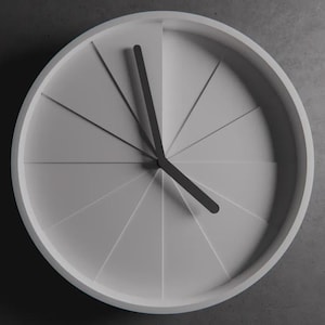 Op de afbeelding: Een minimalistische, moderne wandklok met een lichtgrijze wijzerplaat en een witte rand. De klok heeft een uniek ontwerp met gesegmenteerde secties en zwarte wijzers die de tijd aangeven. Een stijlvol woondecoratie-item.