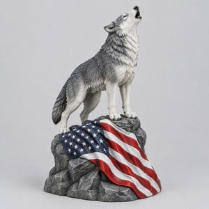 Op de afbeelding: Een grijs en wit wolvenbeeldje dat op een getextureerde grijze rotsformatie staat. De wolf is afgebeeld met de kop omhoog in een huilende houding. Over de rots is een rood, wit en blauwe vlag van de Verenigde Staten gedrapeerd.