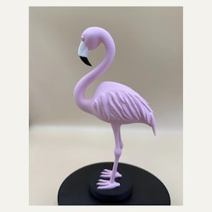 May include: A light pink flamingo figurine standing on a circular black base. The bird features a long, curved neck, textured wing details, and a beak coloured in white and black. The sculpture is rendered in a solid matte finish throughout.