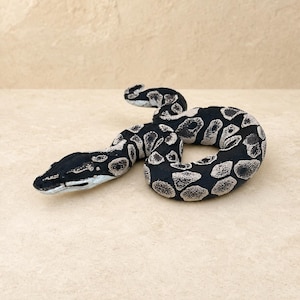 May include: A coiled snake figurine featuring a high-contrast pattern of solid black patches interspersed with textured light gray and white markings. The sculpture depicts a serpent with a distinct head shape and a curved body resting in a loop.