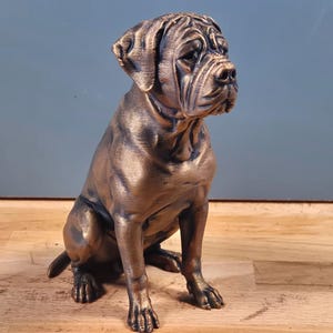 Könnte beinhalten: Eine bronzefarbene Hundefigur in sitzender Position mit faltigem Gesicht und kurzen Ohren. Die Skulptur hat ein metallisches Finish mit dunklen Akzenten in den Vertiefungen, die den muskulösen Körperbau und die aufmerksame Haltung betonen.
