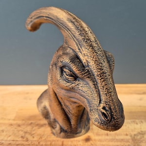 Könnte beinhalten: Eine bronzefarbene Dinosaurier-Büste, die einen Parasaurolophus-Kopf mit einem langen, gebogenen Schädelkamm zeigt. Die Skulptur weist detaillierte Hautstrukturen, tief liegende Augen und ein verwittertes Finish mit dunklen Schattierungen auf.