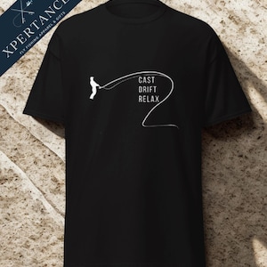 Könnte beinhalten: Schwarzes T-Shirt mit einer weißen Grafik eines Fliegenfischers und den Worten "CAST DRIFT RELAX". Das Shirt hat auch den Text "XPERTANCE FLY FISHING APPAREL & GIFTS" oben links.
