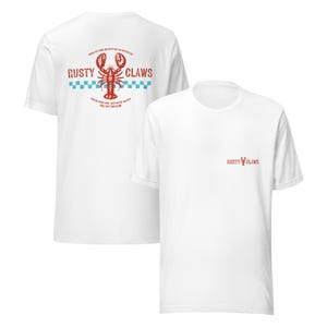 Könnte beinhalten: Weiße T-Shirts mit einem roten Hummer-Motiv und der Aufschrift "Rusty Claws". Die Rückseite des Shirts zeigt ein größeres Design mit Text, einer Hummer-Illustration und einem blau-weißen Karomuster. Die Vorderseite hat ein kleineres Logo.