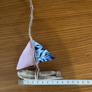以下が含まれることがあります： 流木をベースにした手作りのミニチュア帆船。ピンクと青の模様の布の帆と紐の索具が付いています。ボートの長さは約10cmで、素朴な航海をテーマにしています。
