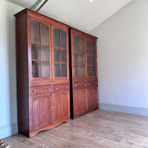 May include: Two tall, matching wooden cabinets with a reddish-brown finish. Each unit features an upper section with glass-paned doors, two small drawers in the middle, and a lower section with solid wooden doors. The cabinets have small round knobs.