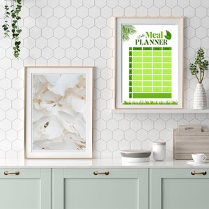 May include: Two framed prints displayed on a white shelf above a mint green cabinet. One print features a close-up of white petals, while the other is a green and white "Daily Meal Planner" with a leaf and grass design.