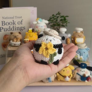 May include: A handmade, crocheted panda bear with black and white details, a small hat, and a yellow flower. The panda is held in a hand, with other crocheted animal figures in the background. The panda is approximately 3 inches in diameter.