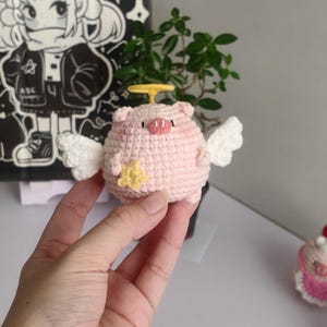 Puede incluir: Un cerdo rosa tejido a mano con alas blancas, un halo amarillo y una estrella amarilla. El cerdo tiene un hocico rosa y ojos negros pequeños. Este artículo caprichoso y artesanal es una pieza decorativa única.