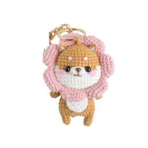 Puede incluir: Llavero de crochet hecho a mano con dise&ntilde;o de perro Shiba Inu. El perro tiene cabeza marr&oacute;n, cara blanca y mejillas rosas, rodeado por un collar en forma de flor rosa. El llavero tiene un anillo de metal dorado.