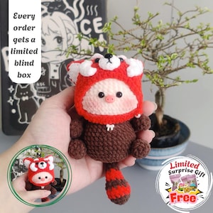 Puede incluir: Muñeco de ganchillo de panda rojo con gorro rojo y blanco, cuerpo marrón y cola a rayas. La imagen incluye texto: "Cada pedido obtiene una caja ciega limitada" y "Regalo sorpresa limitado gratis".